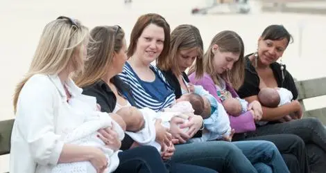A group of 6 women breastfeeding their babies in a line on a park bench next to a river.