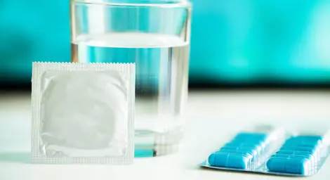 Image of a wrapped condom leaning a glass of water, with blue tablets lying beside on a white table.